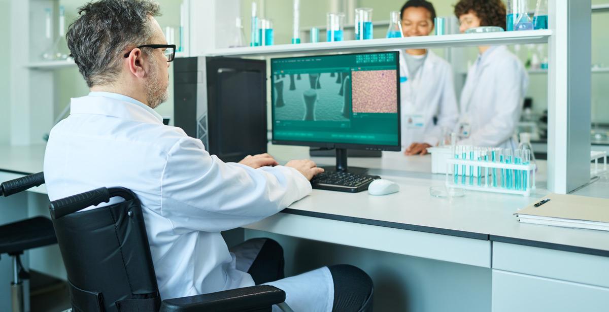 Ricercatore in sedia a rotelle lavora su pc in laboratorio con due colleghe - Foto di pressmaster da Adobe Stock